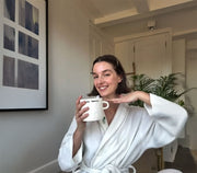 Woman in a white robe holding a mug in a room with a plant and framed picture on the wall.